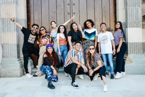 A joyful group of young adults posing in front of a large wooden gate, exuding happiness and diversity.