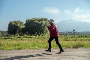 A woman in sportswear jogs along a rural road, surrounded by lush countryside and mountains.