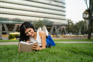 Smiling woman lying on grass reading a notebook in a city park on a sunny day.
