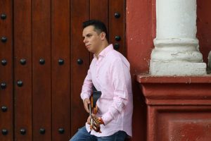 A musician in a pink shirt plays guitar by a rustic wooden doorway, creating a serene street scene.