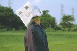 A man stands in a field wearing a newspaper on his head, creating a surreal and artistic vibe.