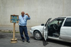 Senior man standing beside his taxi with an open door on a city street.