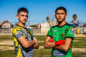 Two teenage soccer players in colorful jerseys pose confidently on an outdoor field under clear blue skies.