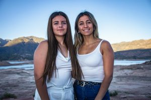 Two young women smile while standing by a beautiful lake with mountains in the background.