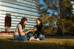 Two young women enjoying a cheerful conversation sitting on grass in an urban park.