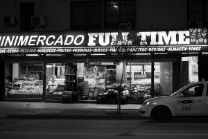 Black and white photo of a minimarket storefront in Córdoba, Argentina, at night with a taxi nearby.