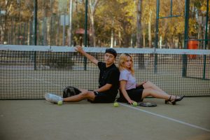 Casual young couple seated back to back on a tennis court with racquets and balls.