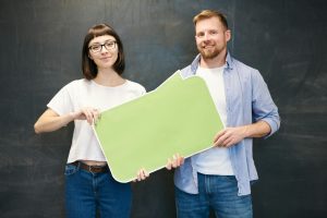 Man and woman with glasses holding a green speech bubble in front of chalkboard.
