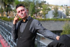 A man with artistic face paint poses confidently outside against a blurred backdrop.