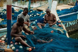 Three African fishermen repairing fishing nets on a traditional boat in Tema, Ghana.
