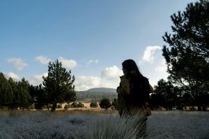 A woman in a yellow jacket hikes through a beautiful mountain landscape with a blue sky backdrop.