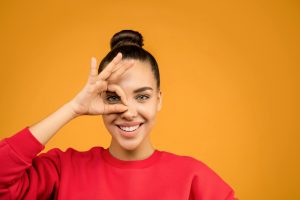 Smiling woman making a playful hand gesture against a vibrant orange background.