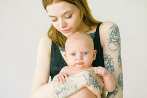 A tattooed mother lovingly holds her baby while smiling gently indoors.
