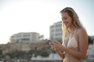 A young woman in a bikini smiles while texting on her smartphone at the beach during summer.