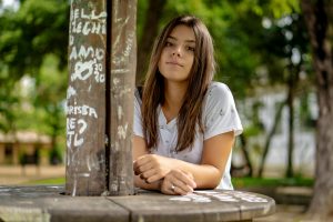 A young woman with long brunette hair poses confidently in an outdoor park setting with graffiti decor.
