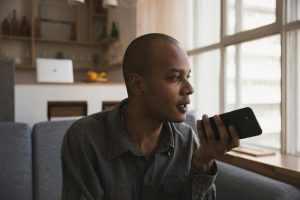 Man holding smartphone and speaking indoors, sitting on sofa by window lighting.