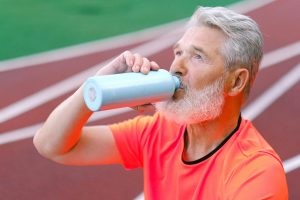 Elderly male with gray beard drinking liquid from thermos while resting after training on blurred background of racetrack