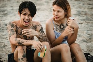Smiling couple with tattoos enjoying drinks while sitting together on the sandy beach.