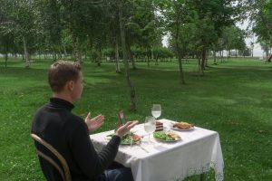 Side view of anonymous male sitting at table with food talking to imaginary date in empty green park