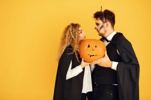Couple in Halloween costumes holding a carved pumpkin, against a yellow background.