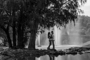 A romantic couple embraces near a scenic bridge and fountain, captured in grayscale.