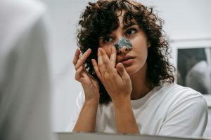 Young female in white t shirt applying blackhead strip on nose standing in front of mirror