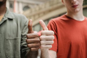 Crop anonymous diverse male friends in casual outfits demonstrating thumbs up gesture while standing together on city street