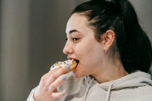 Crop young brunette in comfy hoodie eating fresh appetizing doughnut covered with vanilla icing and sprinkles in light room