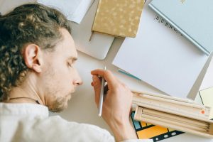 Man demonstrating fatigue falling asleep at desk, surrounded by documents and work materials.