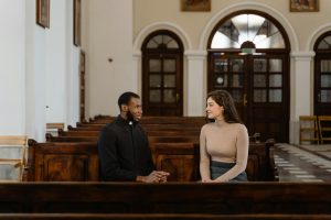 A priest and a woman in conversation sitting in church pews, creating a serene and introspective scene.