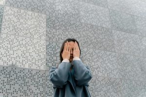 A woman in a stylish blue coat covers her face with hands, standing against a modern perforated metal wall.
