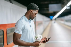 A man in a polo shirt texting on his phone while holding flowers at an empty train station platform.