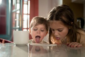 Two adorable siblings playfully licking a table near a mug indoors.