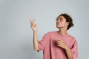Teenage boy with curly hair in pink t-shirt pointing and gesturing against a plain white studio backdrop.