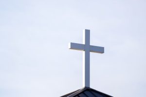 Minimalist image of a white cross on a church roof against a clear blue sky.