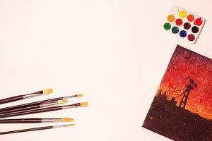 Flat lay of paintbrushes, watercolors, and a vivid canvas artwork with warm tones.