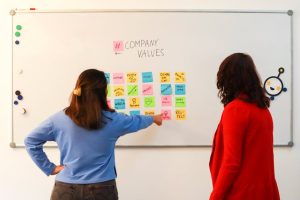 Two women discussing company values on a whiteboard in an office setting, emphasizing teamwork.