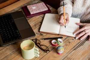 A woman planning her schedule in a notepad beside a laptop and coffee on a wooden desk.