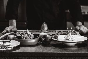 Chef arranges gourmet dishes on a table in a black and white setting.