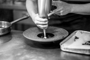 Hands of a chef delicately preparing a dish using a piping tool in a kitchen setting.