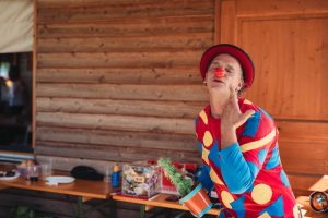 Clown wearing vibrant outfit and red nose, entertaining outside with a toy cactus.