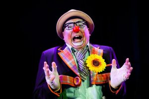 Vibrant clown with red nose and sunflower, expressing joy on stage.