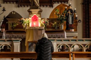 A person praying at a church altar surrounded by religious iconography and flower arrangements.