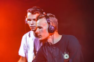 Two DJs perform live with headphones on, creating an immersive music experience under dramatic lighting.