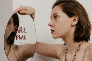 A young woman gazes into a mirror with 'GRL PWR' written on it, emphasizing empowerment.