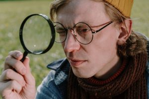 Close-up of a young man with eyeglasses using a magnifying glass in a grassy field.