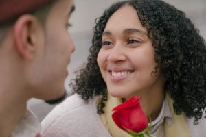 Cheerful Hispanic girlfriend with red flower and crop boyfriend looking at each other while having date on street in city