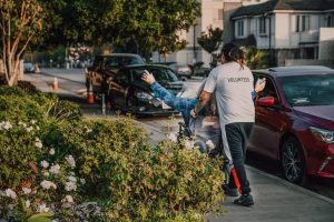 Volunteer assisting a person in a wheelchair on a city street surrounded by greenery and parked cars.