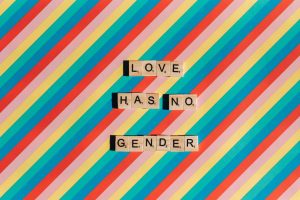 Scrabble tiles spelling 'Love Has No Gender' on a rainbow striped background celebrating Pride.