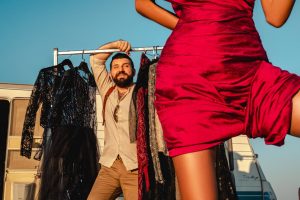 Cheerful man standing near rack with collection of fashionable garments on street with faceless woman and trailer while preparing for video shoot